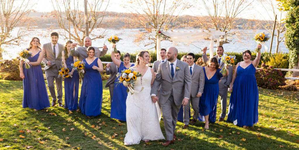 Bride and groom walking at Sea Cliff Manor with bridal party walking behind them. The bride and groom are smiling at each other. 