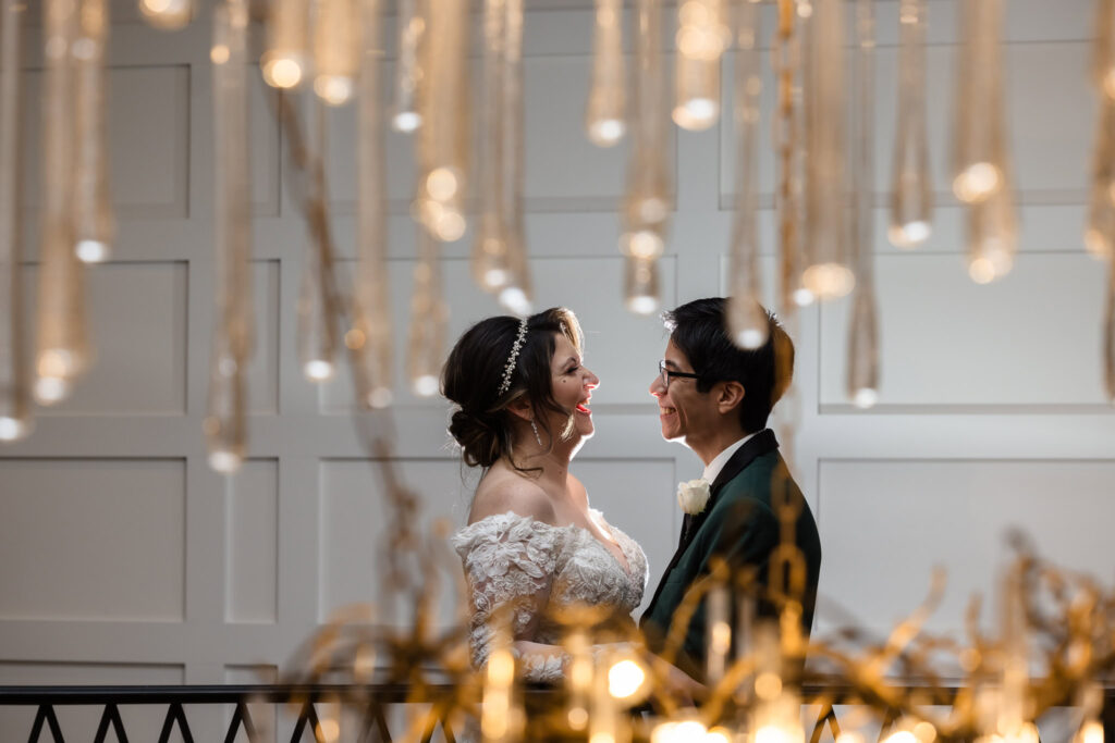 Bride and groom laughing at each other with image taken through a chandelier at Water's Edge