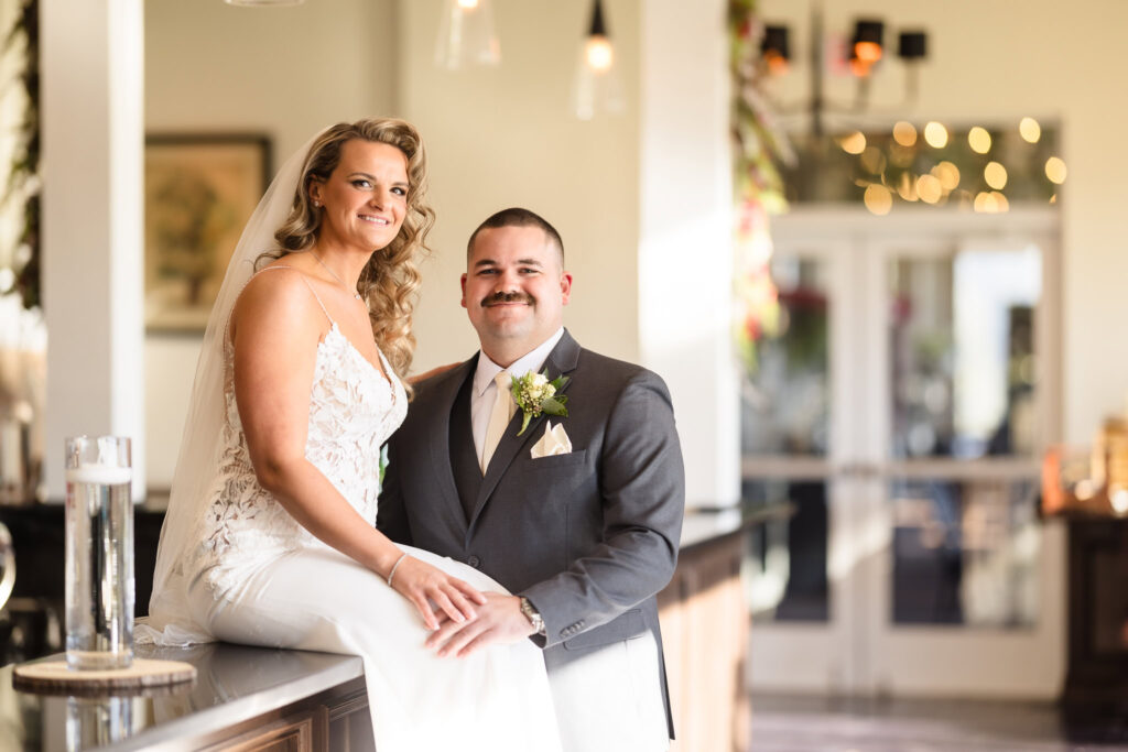 Bride sitting on the bar with groom standing next to her.