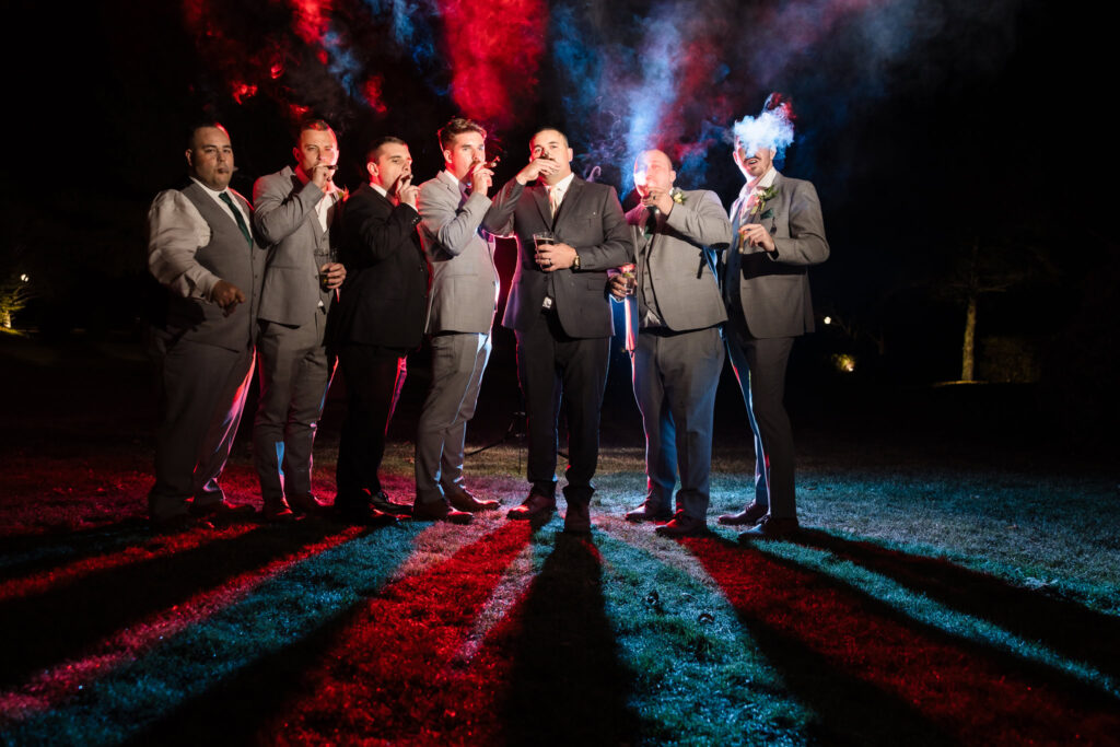 Groom and groomsmen smoking cigars outside. The smoke is backlit and there are red and blue lights illuminating the smoke. 