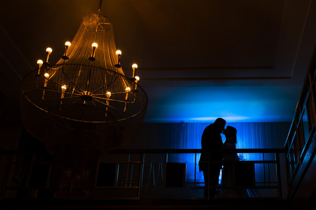 Silhouette of bride and groom kissing with chandelier in foreground.
