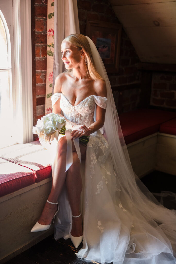 A serene bridal portrait of a bride seated on a window ledge at the Jedediah Hawkins Inn, wearing a white off-shoulder lace wedding dress, a veil, and white high-heeled shoes. She holds a bouquet of white flowers tied with a ribbon. Natural light from the window illuminates her against a backdrop of a brick wall, red cushioned seating, and floral curtains, creating an intimate and elegant atmosphere.