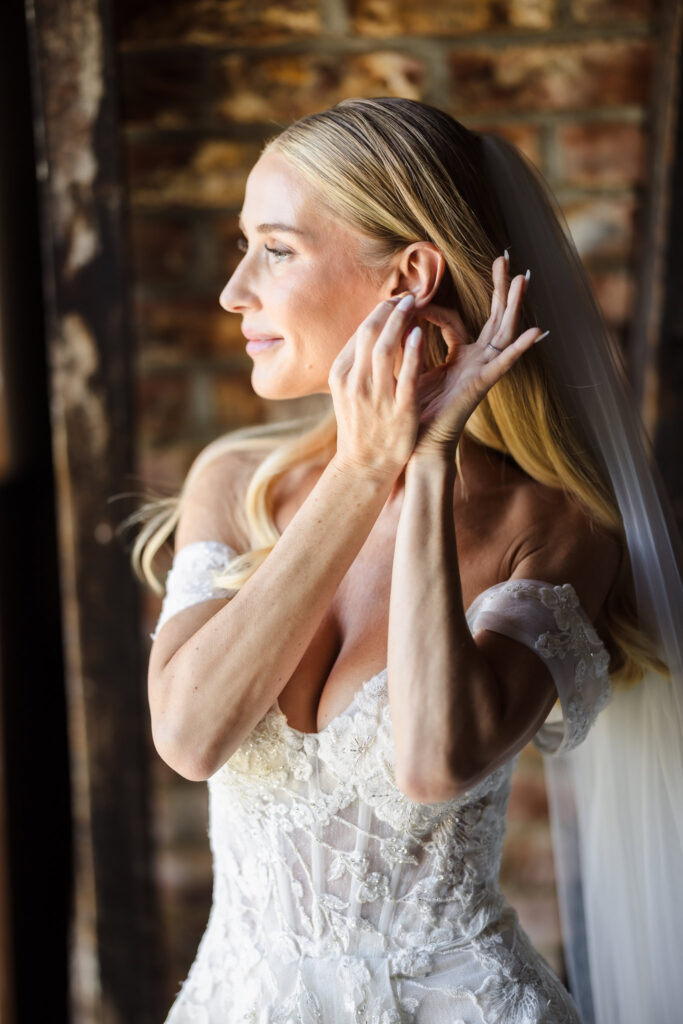 A bride in a white, off-the-shoulder lace wedding dress adjusts her earring, her long blonde hair styled back with a partially visible veil. She is illuminated by natural light from the left, standing against a rustic brick wall that adds warmth and texture to the intimate moment.