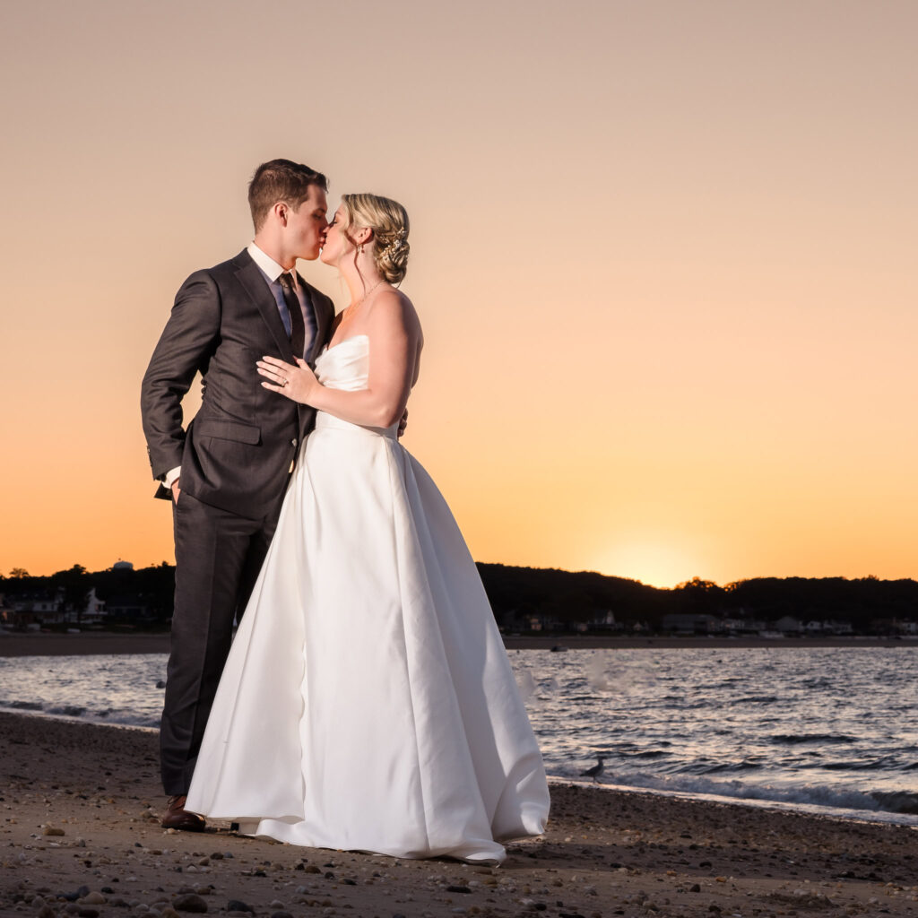 A couple shares a romantic kiss at the Crescent Beach Club at sunset. The person on the left wears a dark suit with a tie, while the person on the right is in a strapless white wedding dress. The warm orange glow of the sunset reflects on the ocean waves, creating a serene and intimate atmosphere.