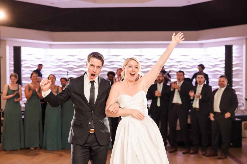 A joyful Crescent Beach Club wedding moment featuring a couple in formal attire. The man, in a black suit and tie, raises his left arm in celebration, while the woman, in a strapless white gown and pearl necklace, raises her right arm. They are surrounded by smiling guests in formal attire, set against a modern white wall with wavy patterns, creating an elegant and festive atmosphere.