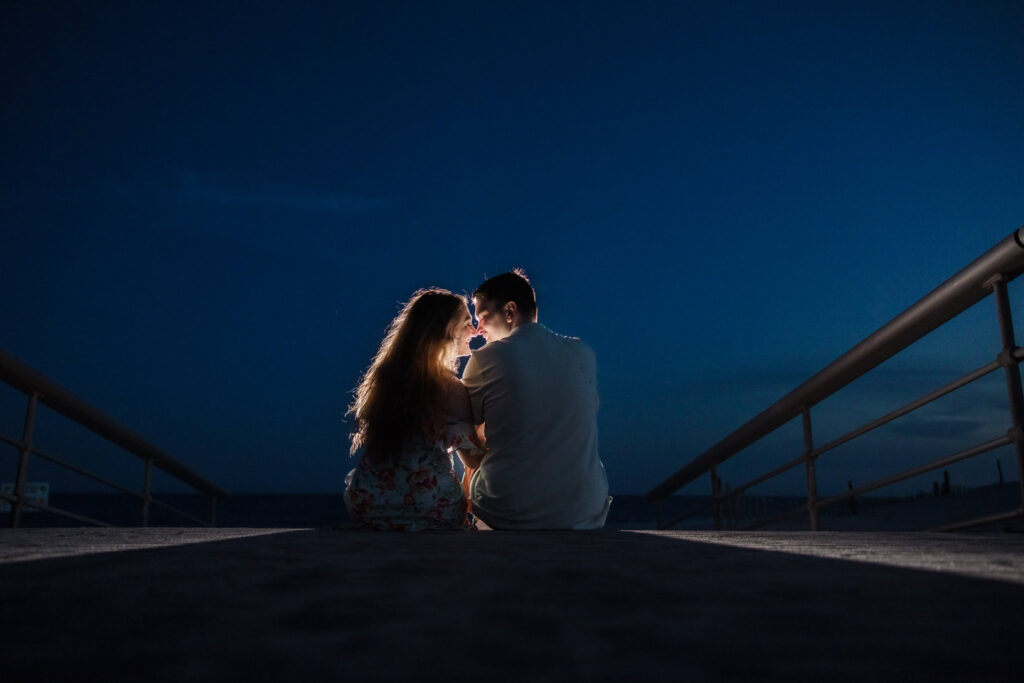 Two individuals sit closely on a paved pathway at night, facing each other in an intimate moment. They are illuminated by soft light, creating a warm contrast against the dark blue sky and shadowy surroundings. Metal railings frame the pathway, leading the viewer's focus to the couple.