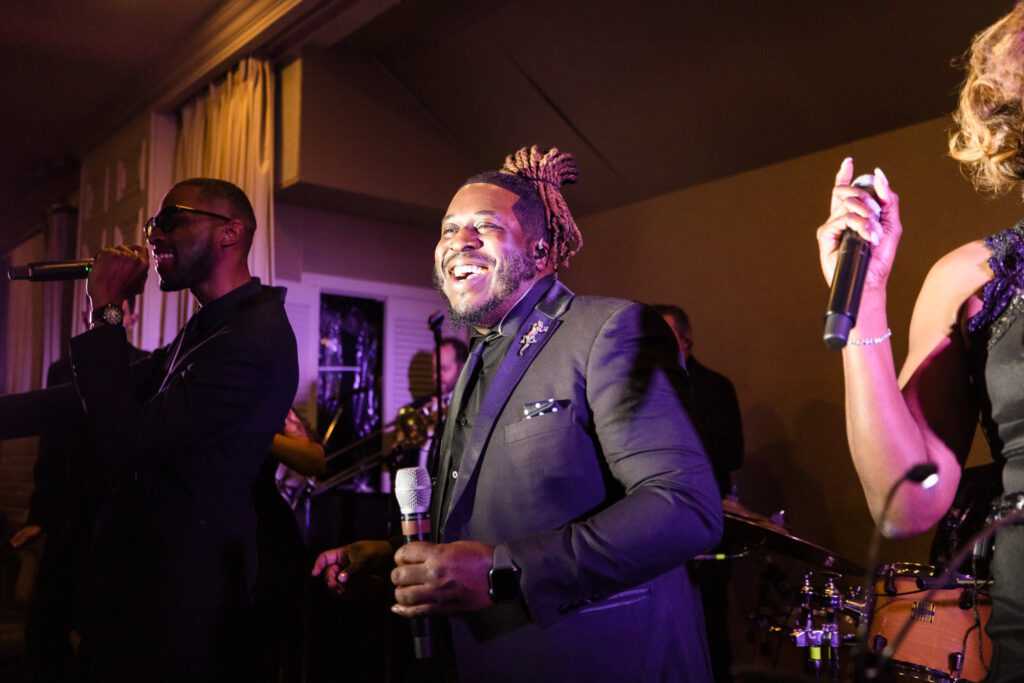 Three performers on stage, each holding a microphone, exude energy and joy during a live performance. The central figure, dressed in a dark suit with a pocket square, smiles broadly, while the person on the left wears sunglasses and sings passionately. Warm lighting highlights the scene, with musical instruments like a drum set visible in the background, creating a lively and celebratory atmosphere.