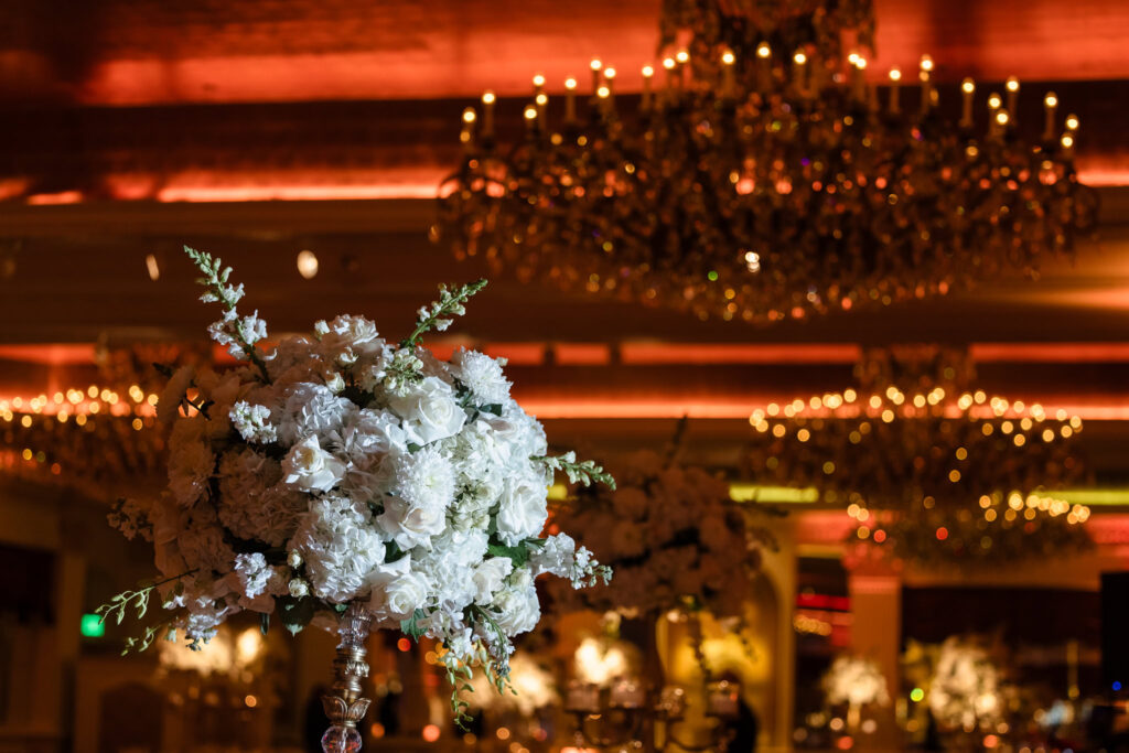 A luxurious floral arrangement at the Garden City Hotel featuring white roses and greenery sits atop a decorative stand in the foreground, sharply in focus. Behind it, ornate chandeliers adorned with glowing lights stretch into the distance, casting a warm, golden ambiance. The blurred background suggests a grand ballroom or upscale event setting.