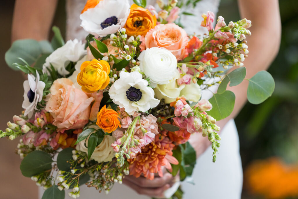 A close-up of a vibrant bridal bouquet featuring white anemones with dark centers, orange ranunculus, peach roses, pale pink snapdragons, white ranunculus, and small white filler flowers, accented with green eucalyptus foliage. The bouquet is held by a person, partially visible in the background.