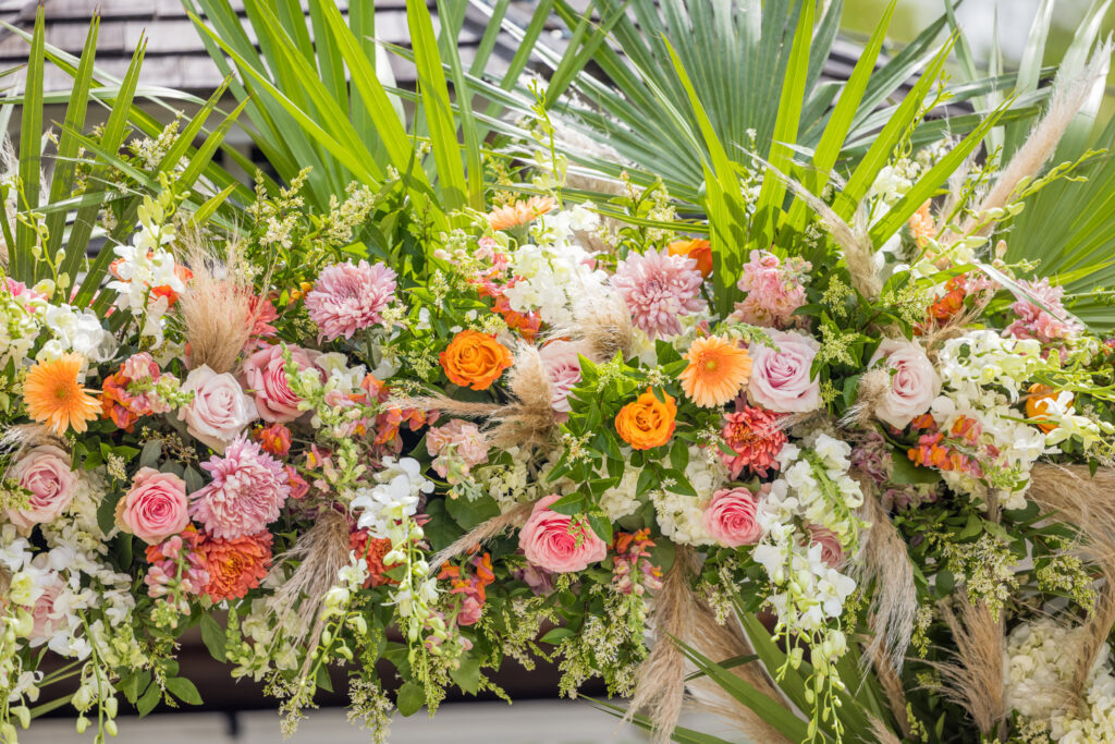 A vibrant floral arrangement featuring pink and orange roses, pink chrysanthemums, white orchids, orange gerbera daisies, and green foliage, including large fan-shaped palm leaves and pampas grass. The lush, asymmetrical design is colorful and textured, ideal for a celebratory NYC wedding.