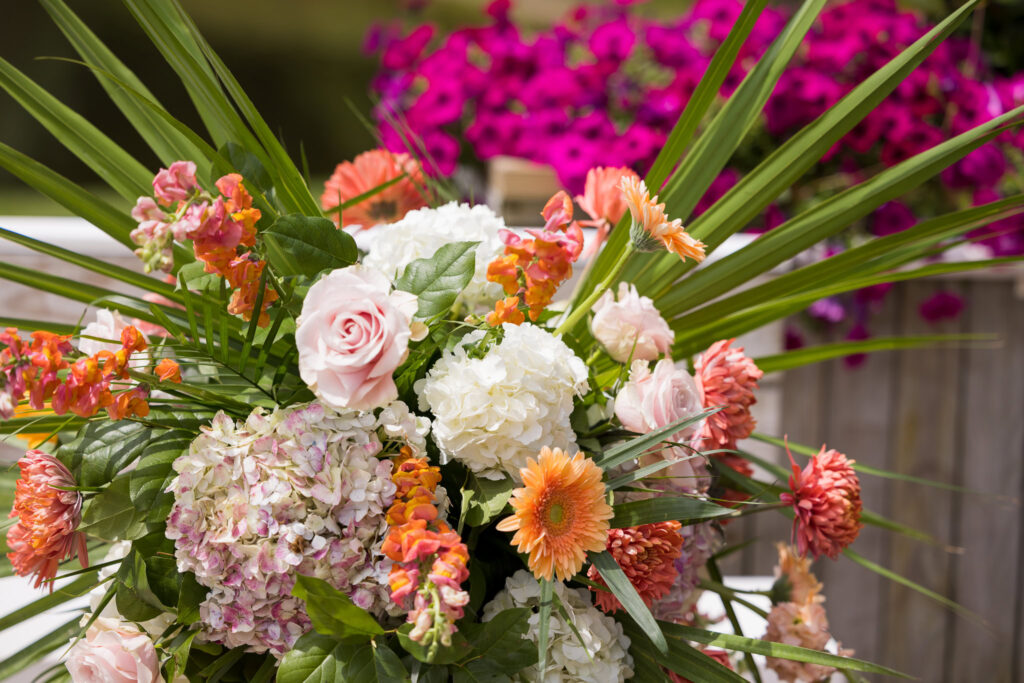 A vibrant floral arrangement featuring light pink roses, white and pink hydrangeas, orange gerbera daisies, orange snapdragons, and pink chrysanthemums, accented with lush green foliage. The flowers are arranged symmetrically, with a blurred background of bright pink blooms, suggesting a festive or celebratory setting.