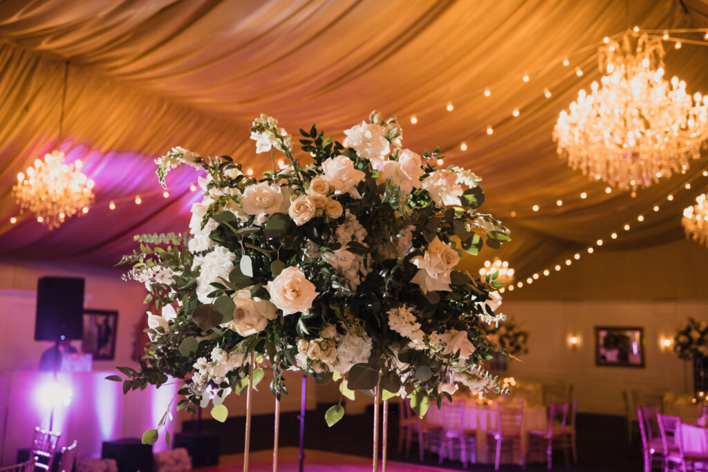 An elegant event space featuring a large floral arrangement of white and cream roses with dark green foliage, elevated on a stand. Warm chandeliers and string lights illuminate the draped gold-toned ceiling, while tables, chairs, and a sound system in the background suggest a formal celebration, likely a wedding or gala.