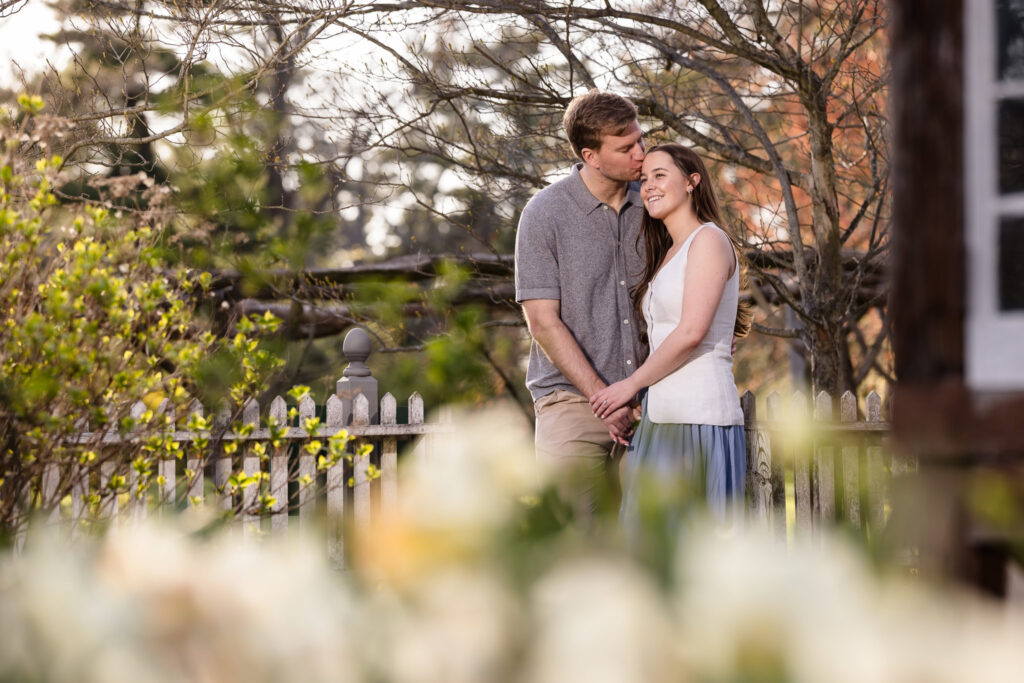 Engaged couple holding hands and smiling in a flowering garden near a white picket fence.
