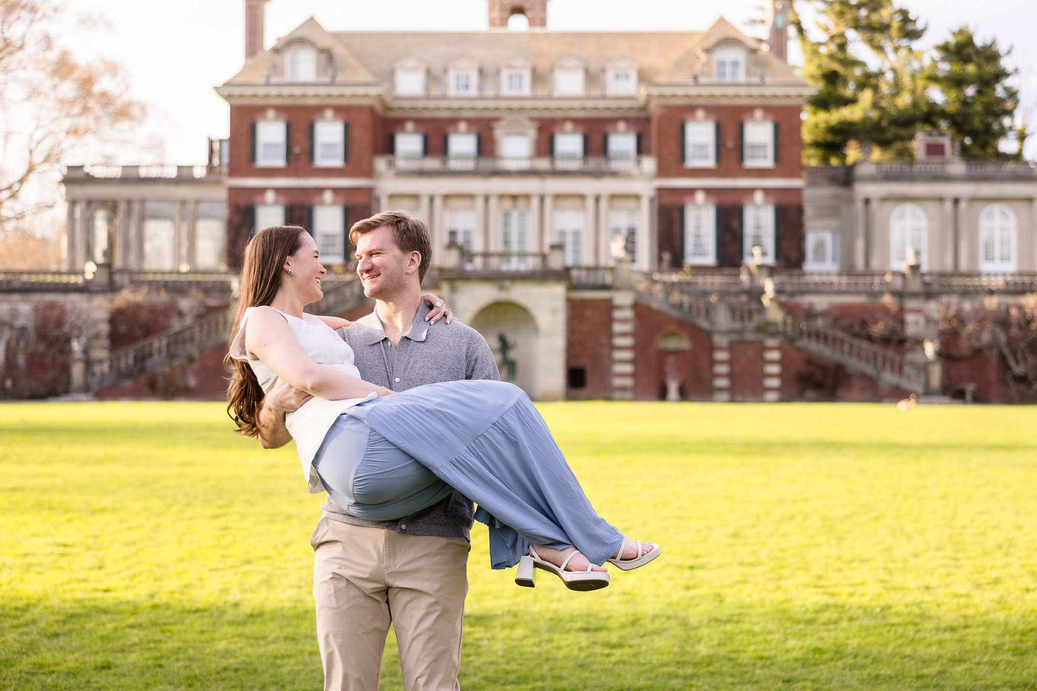 Groom-to-be lifting and smiling at his fiancée in front of the grand estate at Old Westbury Gardens.