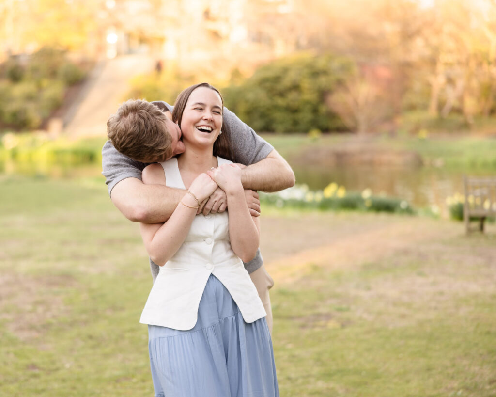 Groom hugging and nuzzling bride from behind as she laughs.