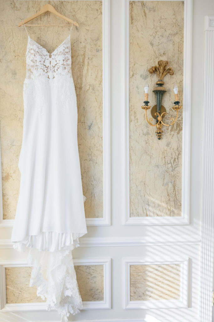 Elegant lace wedding gown hanging on a paneled wall before bridal preparations at a luxury Long Island venue, photographed by Heritage House.