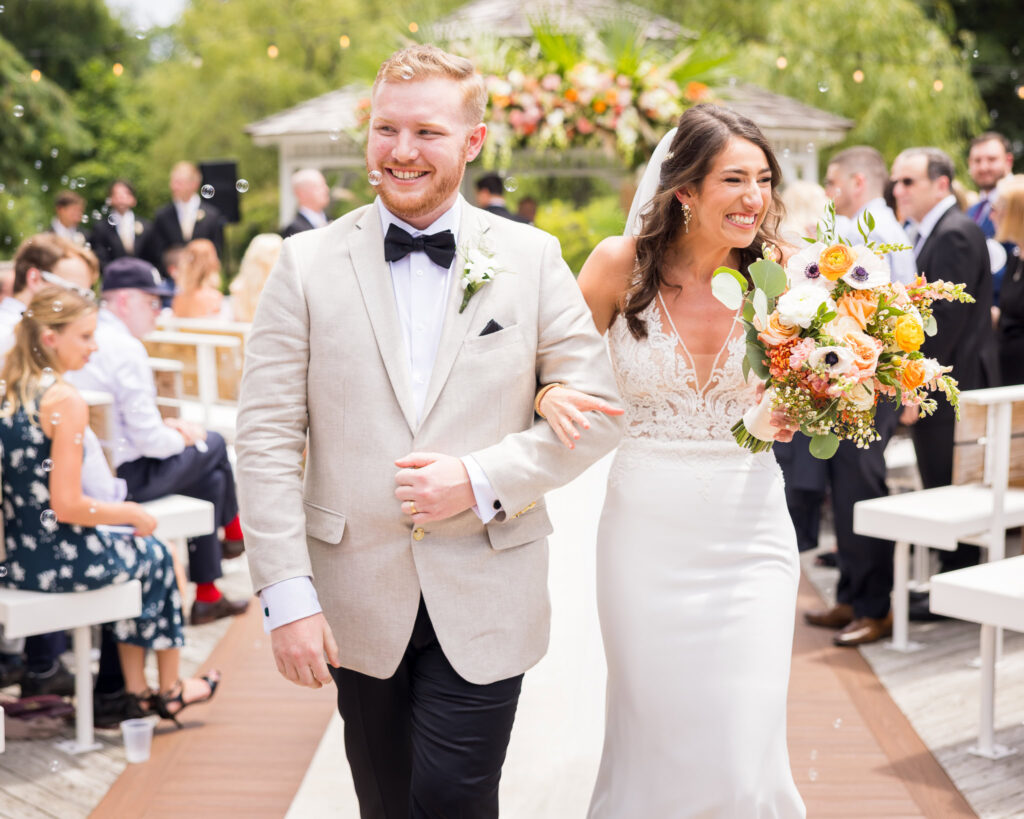 Bride and groom walking back down the aisle, smiling and surrounded by guests and bubbles after their outdoor wedding ceremony in Long Island, captured by Heritage House.