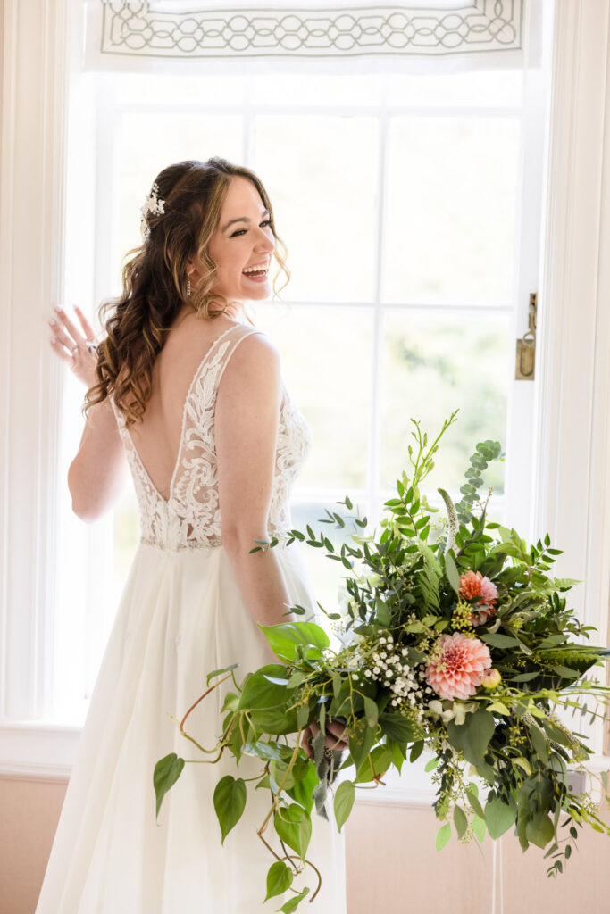 Joyful bride holding a cascading bouquet in front of a bright window, captured in natural light by Heritage House at a Long Island wedding.