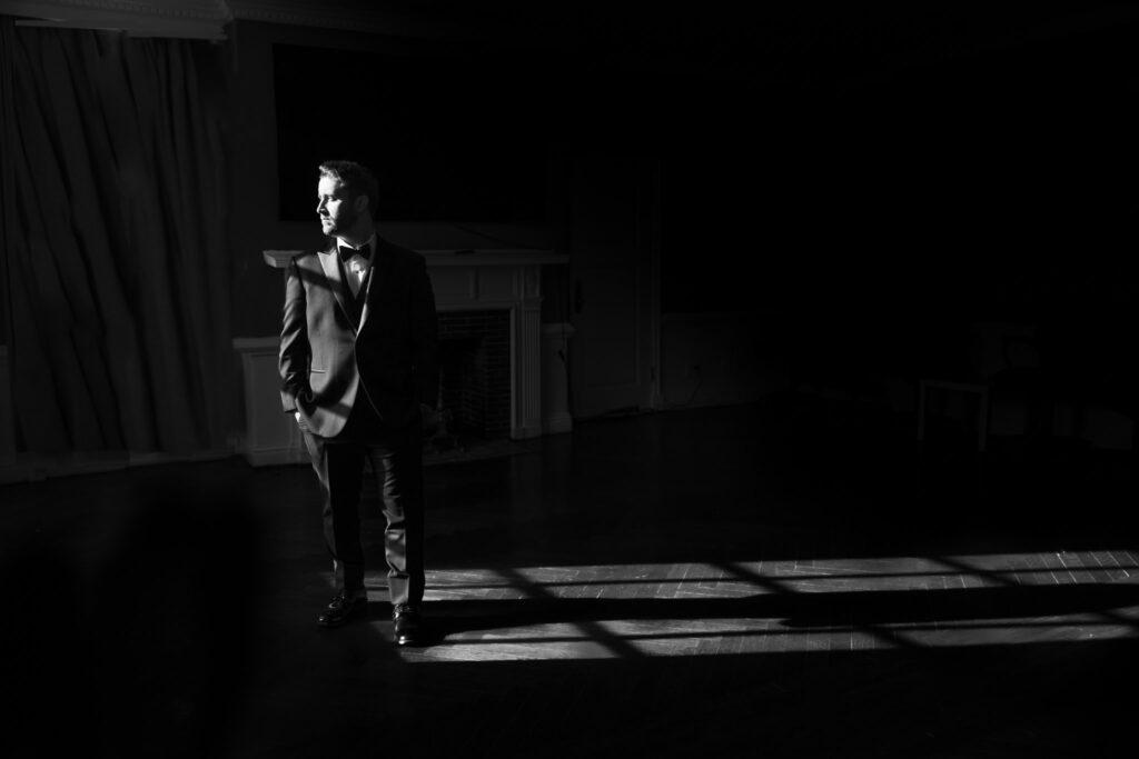 Stylish groom standing in dramatic lighting with deep shadows, captured in black and white by Heritage House for a timeless and cinematic wedding portrait.