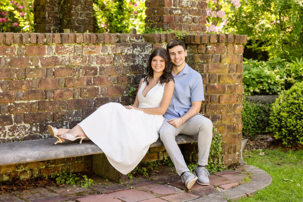 Morgan and Michael sit close together on a stone bench against a weathered brick wall in the gardens at Planting Fields Arboretum. Surrounded by soft greenery and dappled light, they smile warmly, sharing a moment of relaxed joy and quiet closeness.