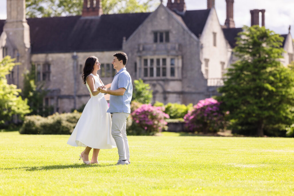 An engaged couple shares a joyful, light-filled moment on the lawn at Planting Fields Arboretum, holding hands in front of the grand stone façade of Coe Hall. Her white dress floats as she steps forward, surrounded by blooming spring foliage under the afternoon sun.