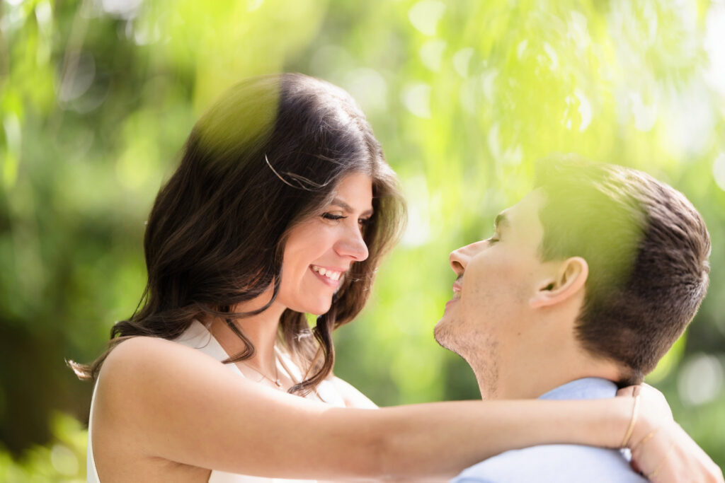 Framed by soft greenery, Morgan smiles as she looks into Michael’s eyes, her arms resting around his shoulders in a close, joyful embrace. Light filters through the leaves above, casting a golden glow on their shared moment.