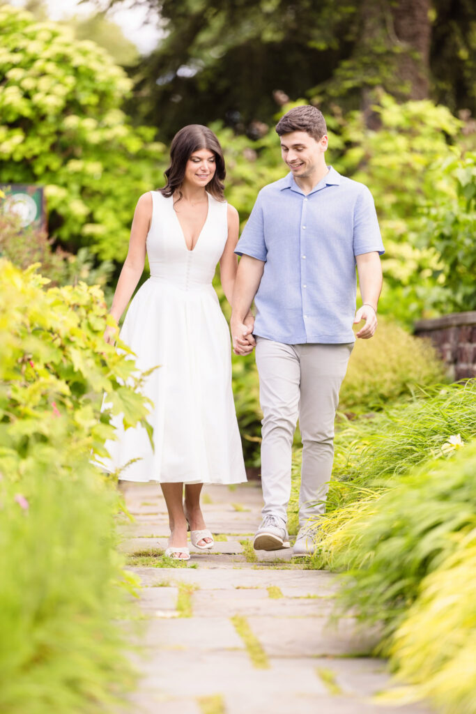 Morgan and Michael walk hand in hand down a stone garden path at Planting Fields Arboretum, surrounded by lush greenery and soft spring textures. Their smiles are relaxed and natural, framed by vibrant foliage in full seasonal bloom.