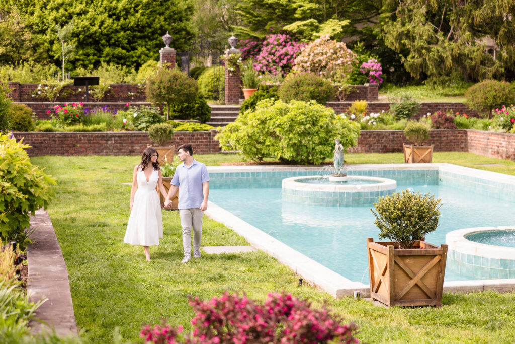 Morgan and Michael walk hand in hand alongside a turquoise reflecting pool in the Italian Garden at Planting Fields Arboretum. Surrounded by manicured hedges, terraced blooms, and classical statuary, the couple’s connection feels serene and timeless.
