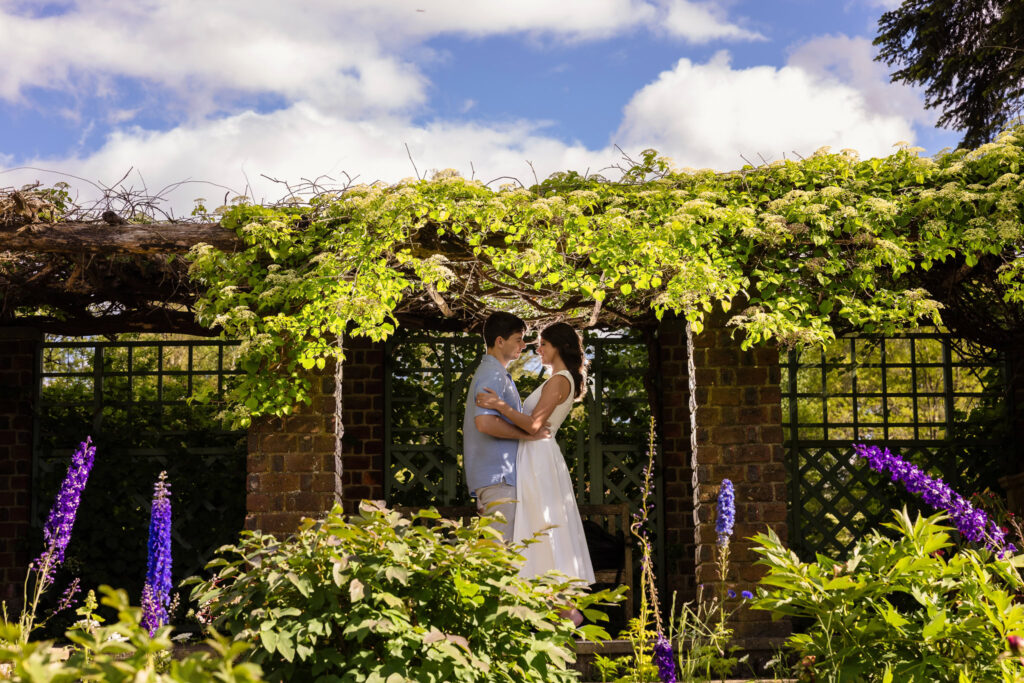 Beneath a vine-covered stone trellis at Planting Fields Arboretum, Morgan and Michael share a quiet embrace, surrounded by flowering greenery and framed by the symmetry of garden architecture. Purple blossoms and soft afternoon light bring the moment to life.