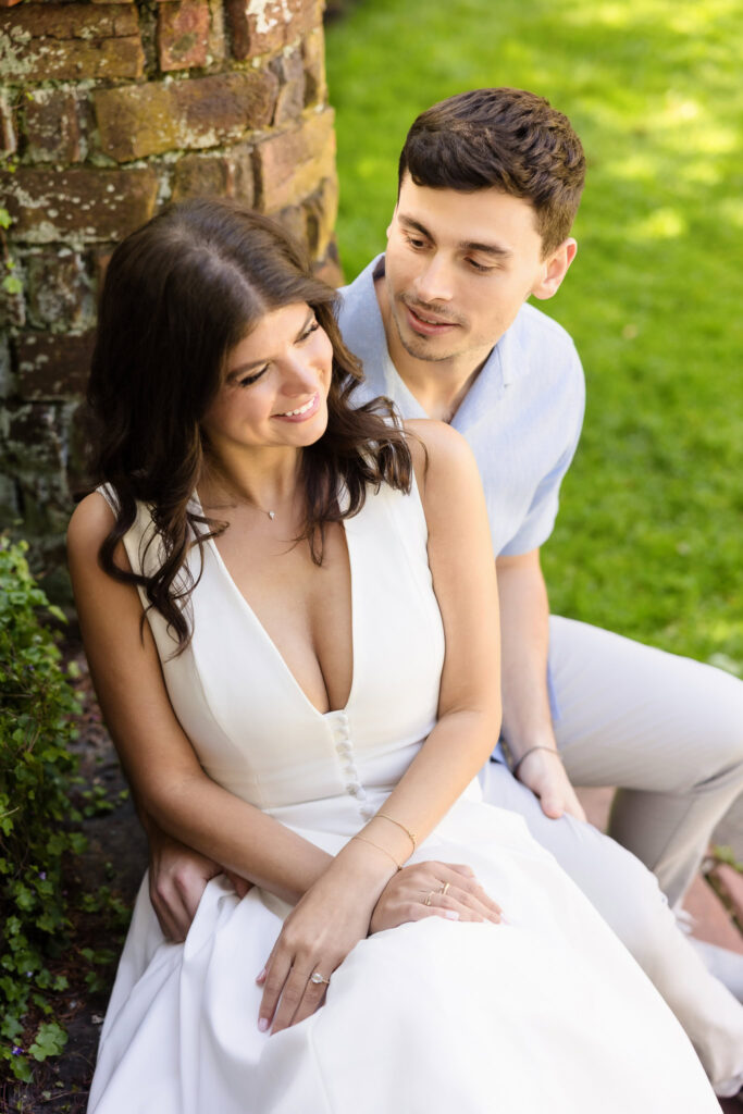 Seated together near a brick garden wall at Planting Fields Arboretum, Morgan smiles as she leans slightly toward Michael, who gazes at her lovingly. Her hands rest delicately in her lap, framed by soft natural light and lush garden greens.