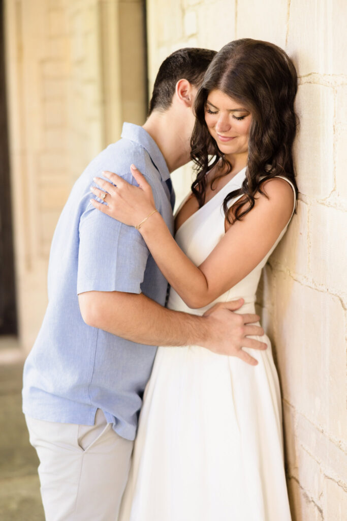 A bride-to-be smiles gently with her eyes closed as her fiancé leans in tenderly, framed against the sunlit stone wall of Coe Hall at Planting Fields Arboretum. Her hand rests softly on his shoulder, revealing her engagement ring in a moment of quiet connection.