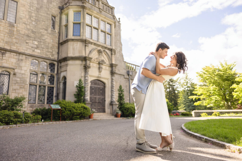 A groom-to-be gently dips his fiancée in front of the ornate stone façade of Coe Hall at Planting Fields Arboretum. The couple smiles into each other’s eyes, bathed in soft afternoon light, surrounded by timeless architecture and manicured gardens.
