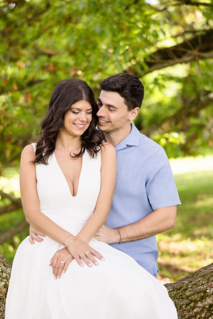 The couple sits close beneath a canopy of trees at Planting Fields Arboretum, framed by soft light and layered greenery. Michael gently embraces Morgan from behind as they smile together, her hands resting gracefully in her lap with her ring delicately visible.