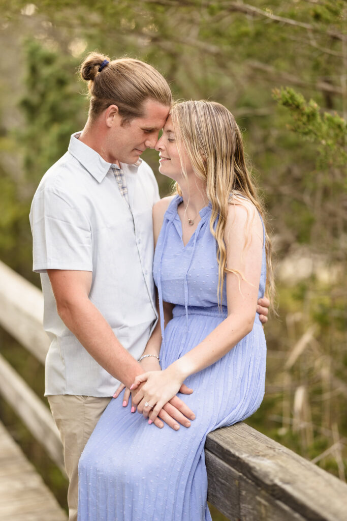 Rachel and Joe share a tender forehead-to-forehead moment while seated on a rustic wooden railing along the Fire Island boardwalk, surrounded by soft greenery and early spring light, her engagement ring gently nestled between their hands.