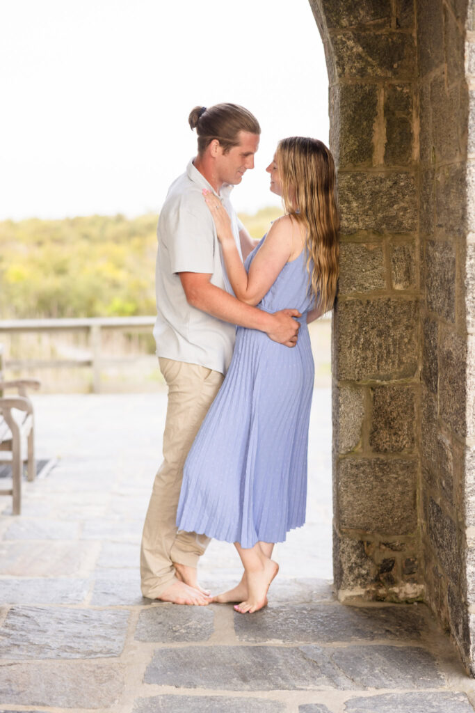 Rachel and Joe stand barefoot beneath a stone archway at Robert Moses State Park, sharing a quiet moment as she lifts her heel with a smile, surrounded by soft light and the natural textures of the historic beach pavilion.