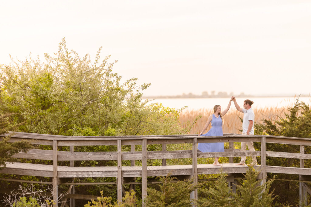 Rachel and Joe twirl barefoot on a wooden boardwalk surrounded by lush greenery and golden reeds, their joy framed by the soft, coastal light of early evening at Robert Moses State Park.