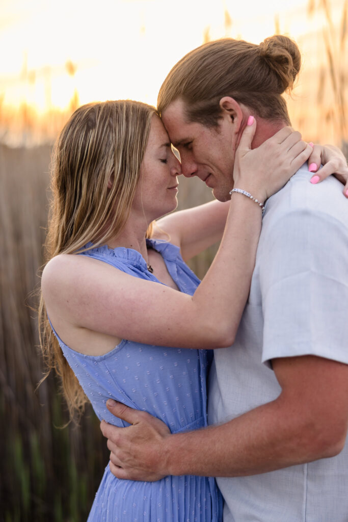 Rachel and Joe share an intimate embrace at sunset in the tall dune grasses of Fire Island, their foreheads gently touching as the golden light surrounds them in warmth and quiet connection.