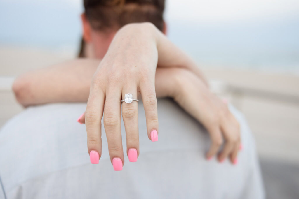 A close-up of Rachel’s hand wrapped around Joe’s shoulders, showcasing her radiant cushion-cut engagement ring and soft pink nails, captured along the boardwalk at Robert Moses State Park.
