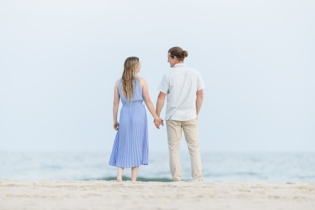 Rachel and Joe stand barefoot at the edge of the beach, hand in hand, gazing out at the horizon over the Atlantic Ocean — a serene, forward-looking portrait from their Fire Island engagement session.