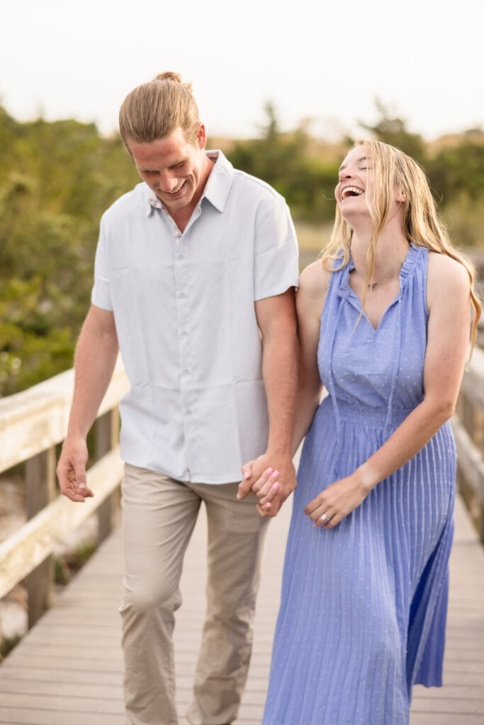 Rachel and Joe walk hand-in-hand along the boardwalk at Robert Moses State Park, sharing a carefree laugh as golden evening light bathes the path behind them — a playful moment that captures the spirit of their spring engagement session.