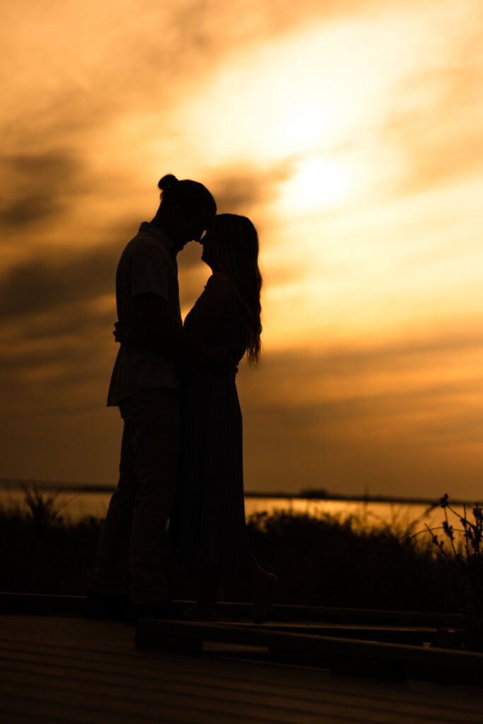 Rachel and Joe share a quiet embrace in silhouette against a glowing amber sky at sunset, standing on the boardwalk at Fire Island with the golden light casting a romantic, timeless mood over their beach engagement session.