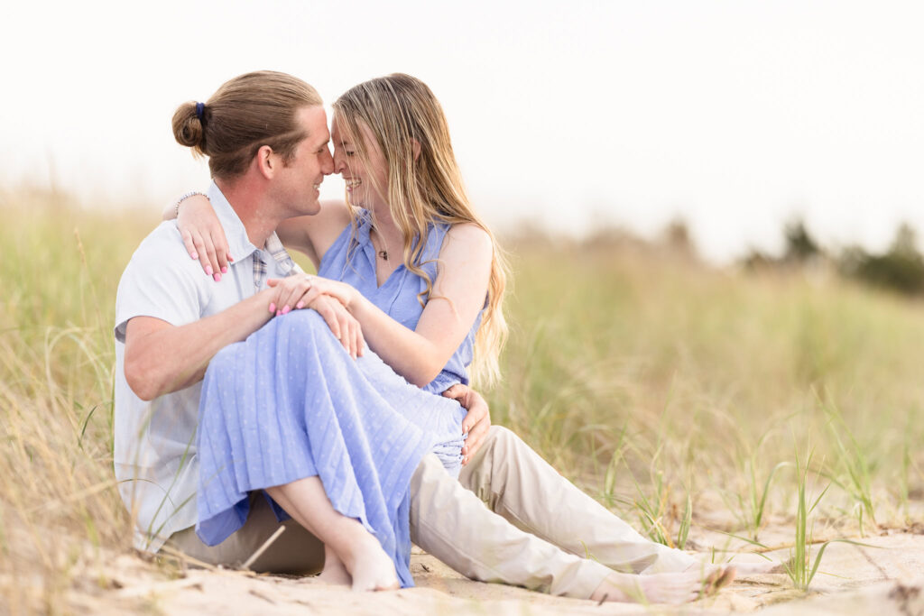 Rachel and Joe sit barefoot in the sand dunes of Fire Island, wrapped in each other’s arms and laughter, framed by soft grasses and golden evening light during their romantic beach engagement session.