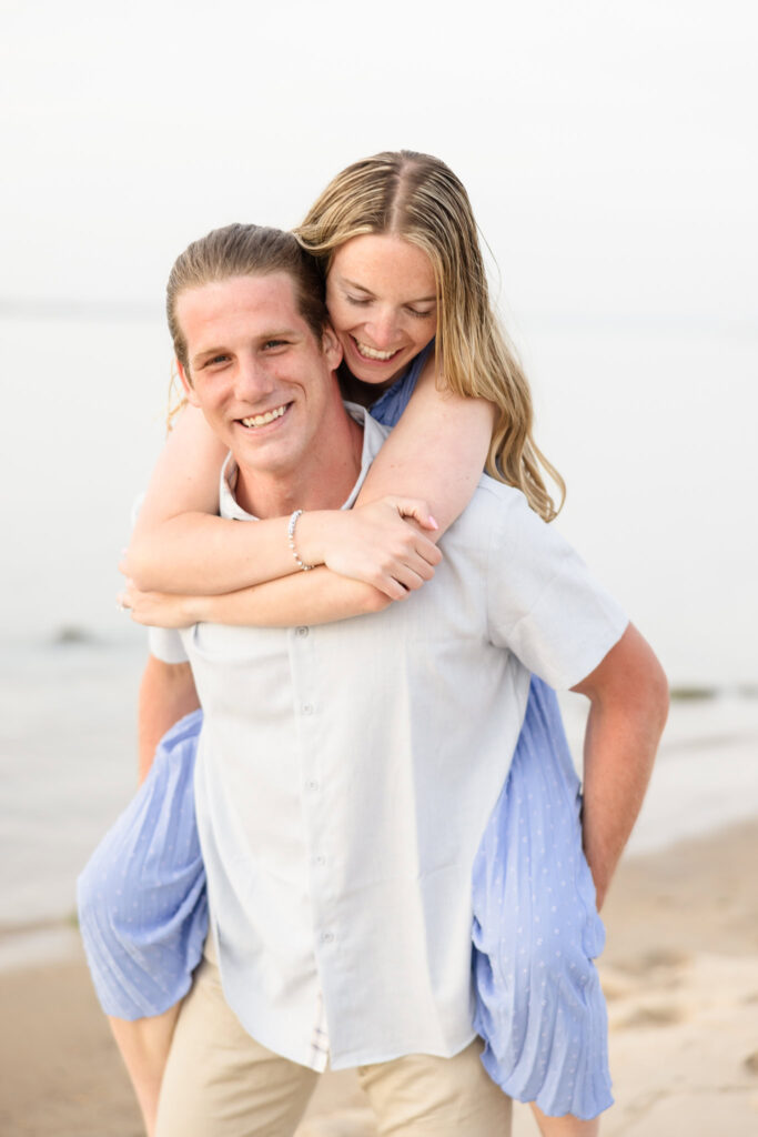 Rachel laughs as Joe carries her on his back along the beach at Robert Moses State Park, her arms wrapped around him as the soft shoreline stretches into the distance — a playful, love-filled moment from their spring engagement session.