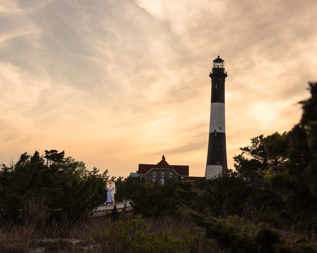 Rachel and Joe share a quiet embrace at sunset beneath the Fire Island Lighthouse, framed by warm skies and coastal evergreens — a cinematic moment that captures the historic charm and emotional depth of their Robert Moses engagement session.