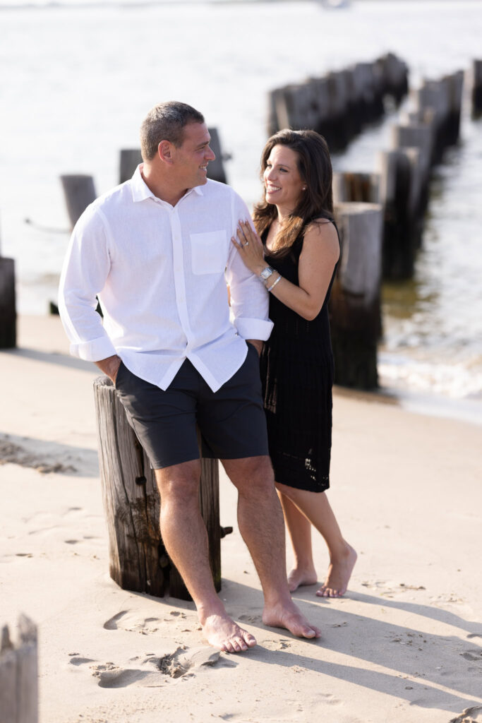 Nina and Scott smiling at each other by the weathered wooden pilings at Breezy Point, with gentle ocean light and a relaxed coastal vibe.