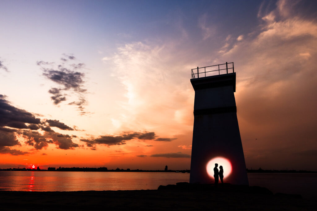 Silhouetted couple standing beneath the Breezy Point lighthouse at sunset, with a glowing circle of light surrounding them and vivid sky reflections over the water.
