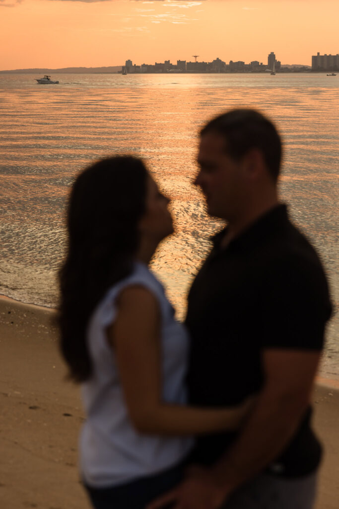 Out-of-focus portrait of Nina and Scott at Breezy Point, with golden sunset reflecting across the water and the Coney Island skyline in the distance.