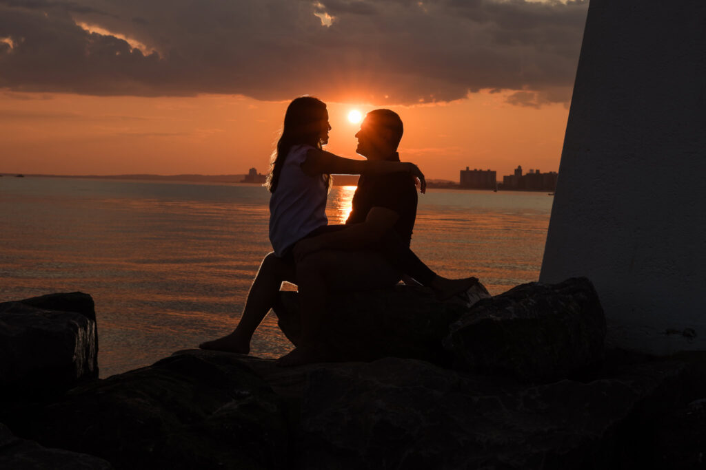 Silhouette of Nina and Scott sitting on the rocks at Breezy Point during their engagement session, with the sun setting behind them over calm water.