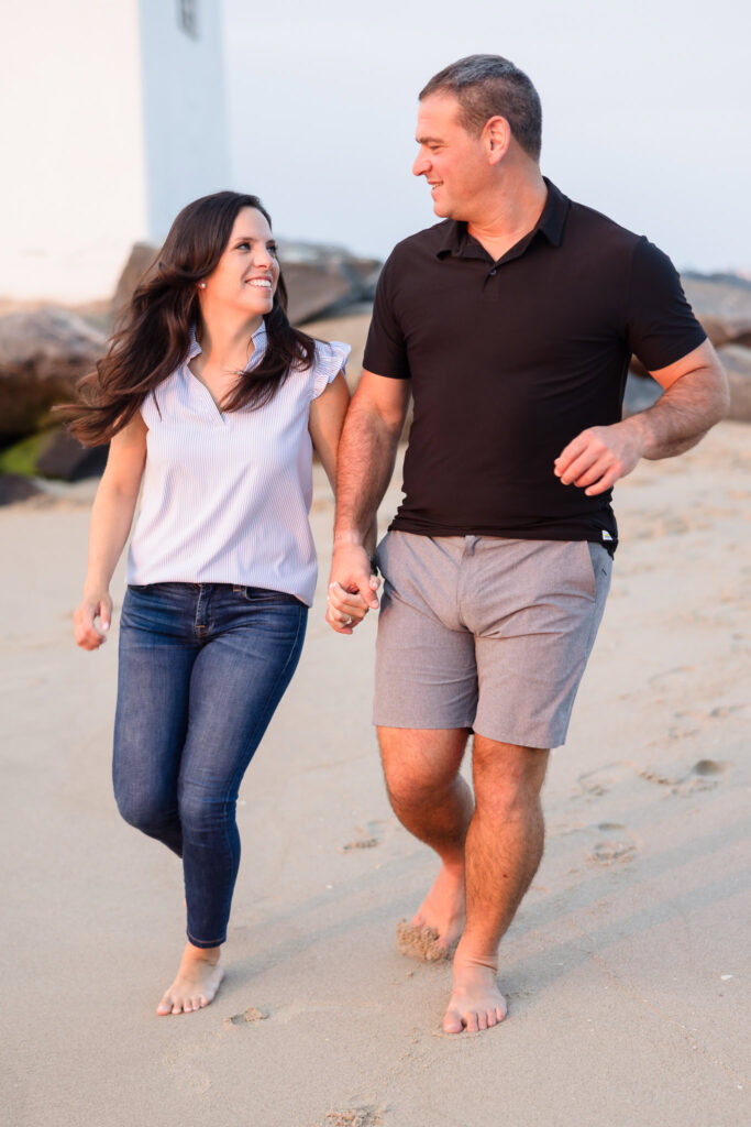 Nina and Scott walking hand-in-hand along the sand at Breezy Point during their engagement session, sharing a warm look in the early evening glow.