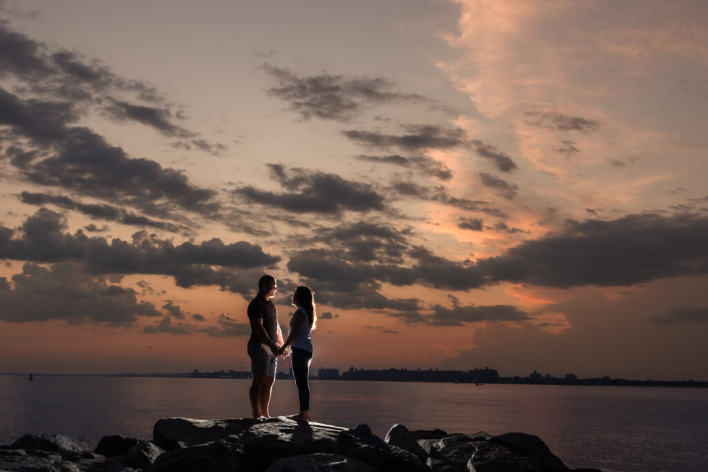 Couple holding hands on rocks at Breezy Point Beach with a dramatic sunset sky over the Manhattan skyline